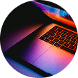 Częściowo widoczny, kolorowo podświetlony laptop sfotografowany z góry, na czarnym tle w okrągłej ramce.